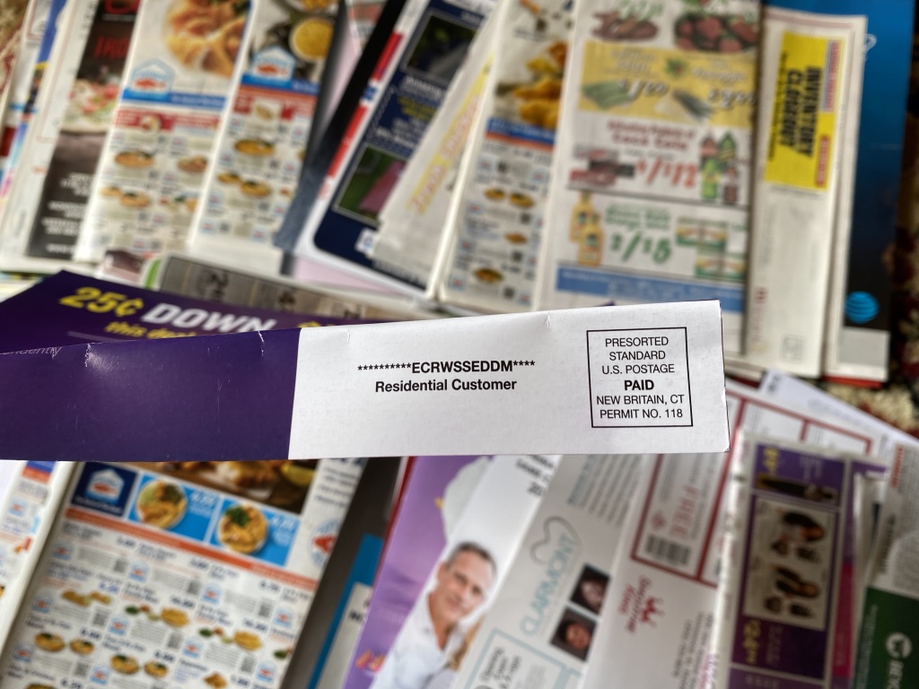 An EDDM flyer addressed to Residential Customer, printed with the ECRWSSEDDM marking and a Presorted Standard permit, lying on a pile of grocery and pizza ads